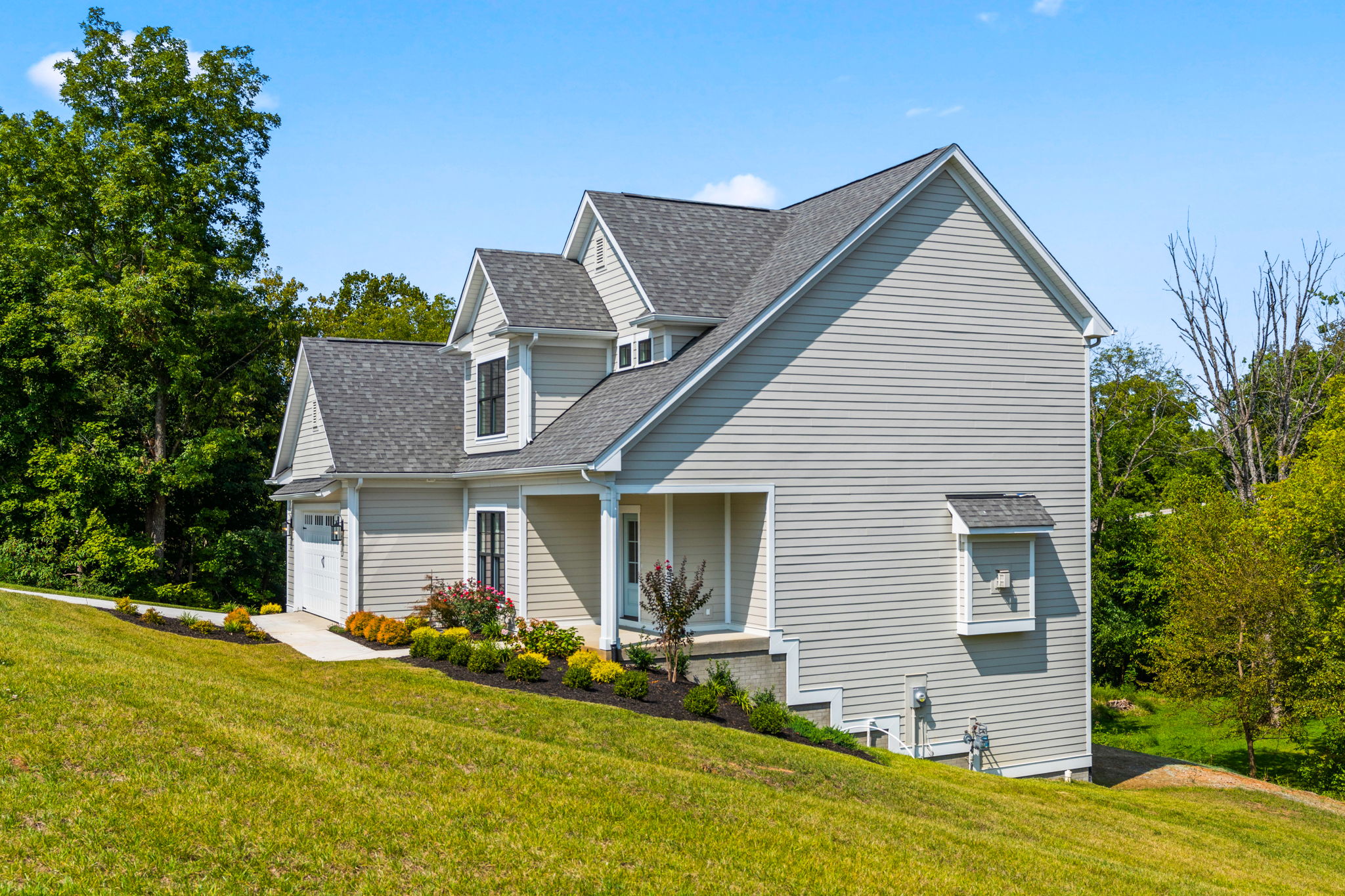 Front view of a two-story beige house with multiple gabled roofs and white trim on a sunny day, with green lawn and trees nearby.