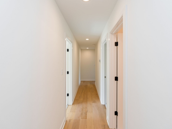 Long, bright hallway with white walls, multiple white doors on both sides, and a light wood floor. Recessed ceiling lights visible down the corridor.