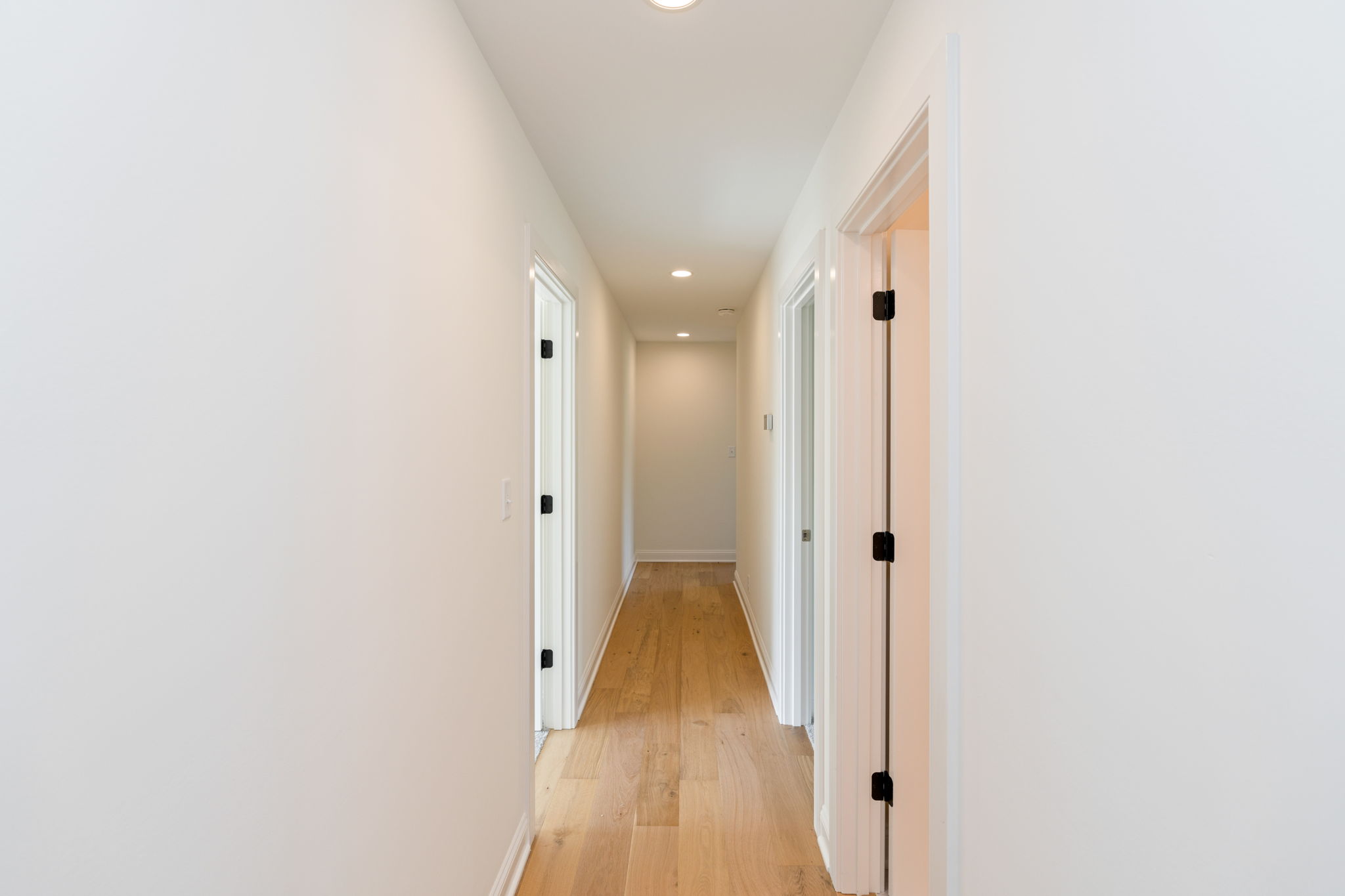 Long, bright hallway with white walls, multiple white doors on both sides, and a light wood floor. Recessed ceiling lights visible down the corridor.