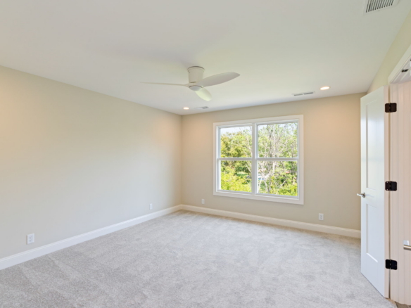 Empty beige bedroom with a white ceiling fan, a large window showing trees outside, and a white door ajar.
