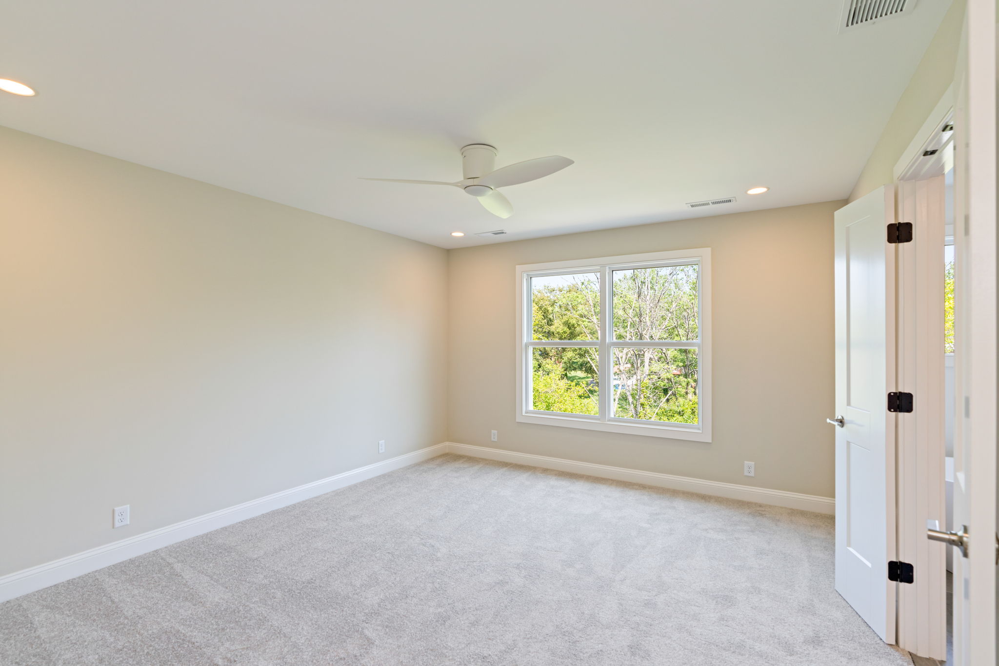 Empty beige bedroom with a white ceiling fan, a large window showing trees outside, and a white door ajar.