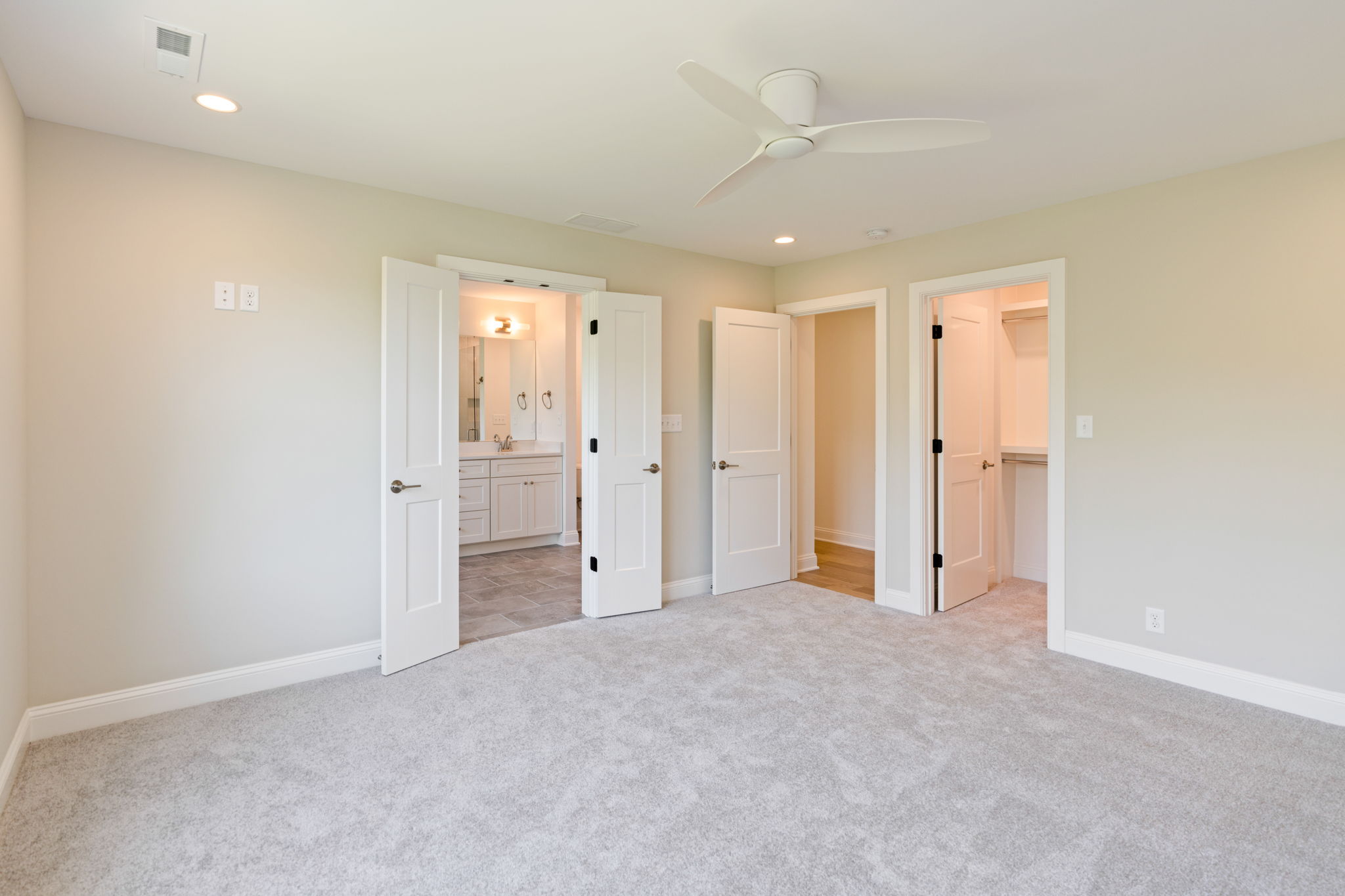 Empty bedroom with beige walls, light carpet, and white doors; open door reveals a bathroom with vanity and mirror.