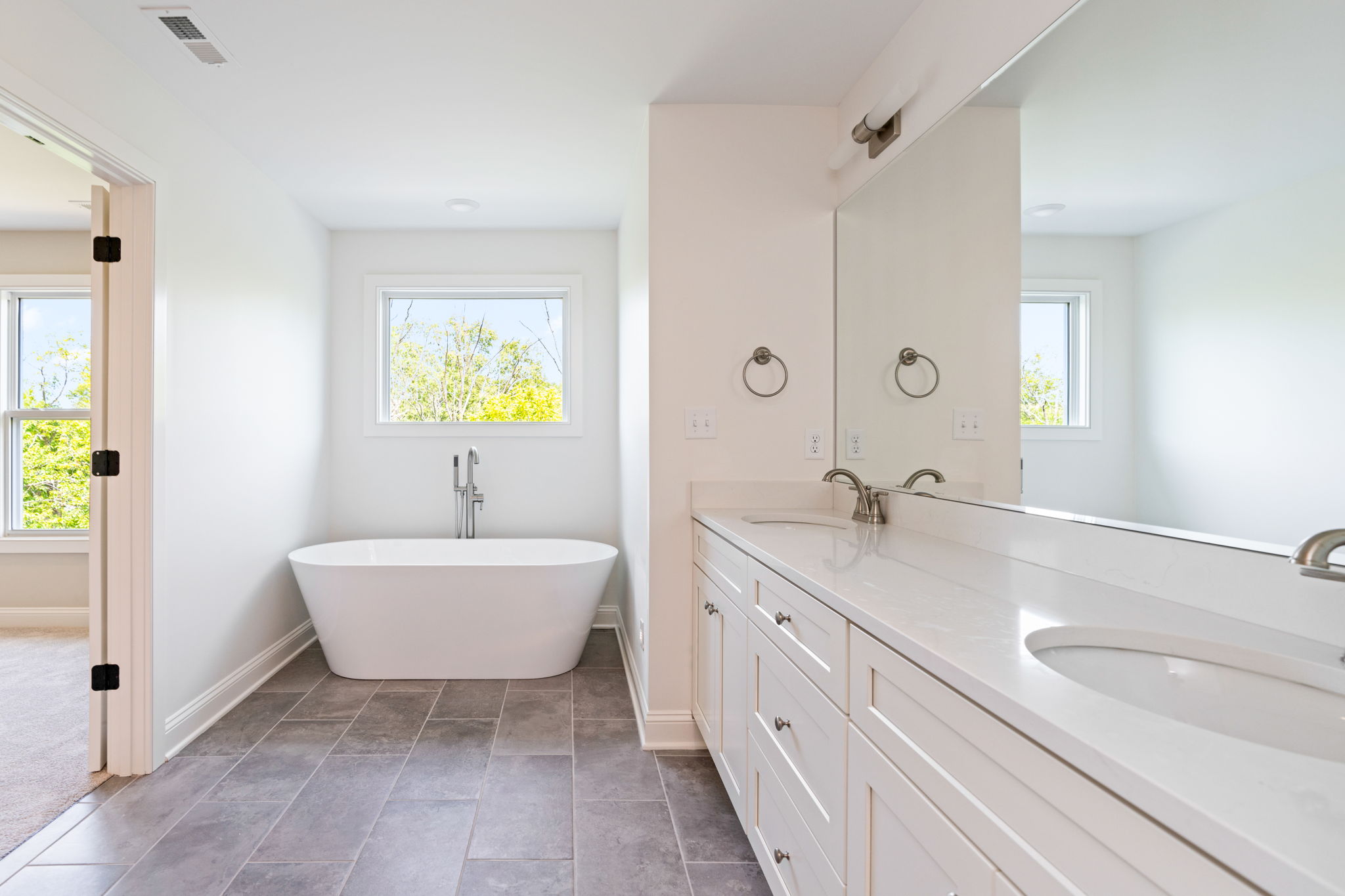 Modern white bathroom with a freestanding oval tub beneath a window and a long double-sink vanity with chrome faucets on the right.