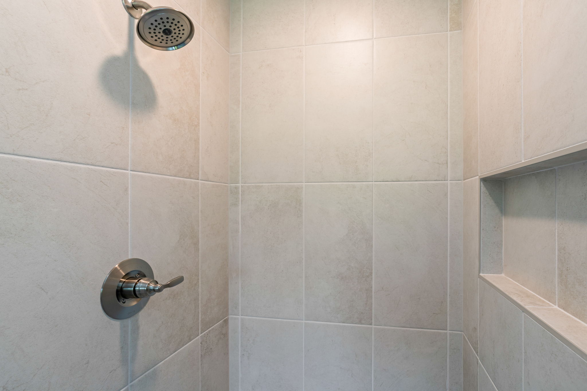 Shower with a round metal rainfall-style head and a lever handle on tiled walls, plus built-in corner shelf to the right.