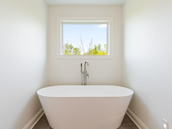 White freestanding bathtub centered in a small bright bathroom with a window showing blue sky and trees outside.