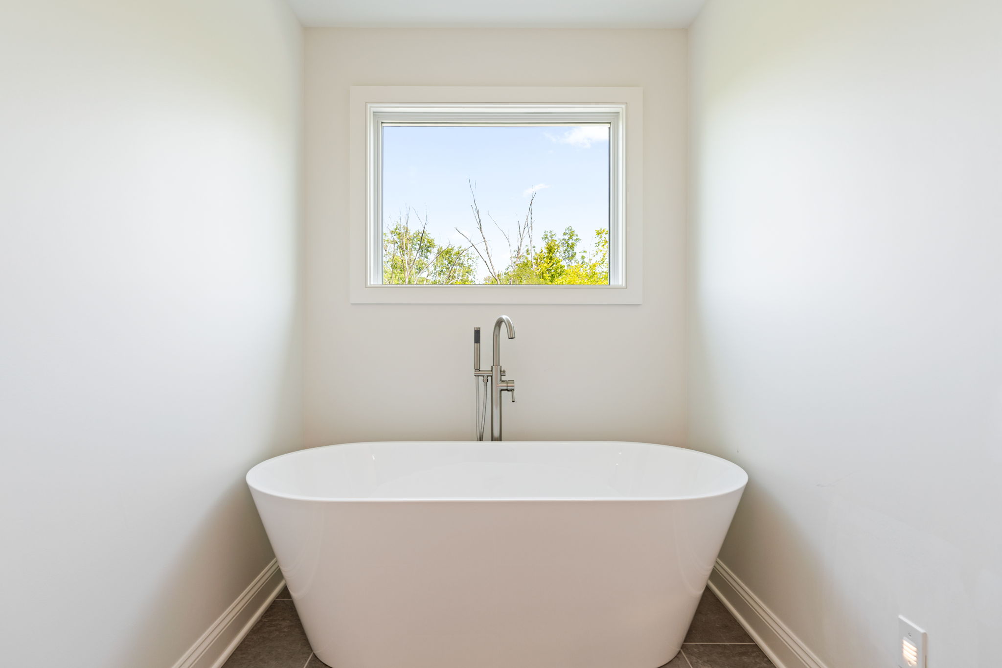 White freestanding bathtub centered in a small bright bathroom with a window showing blue sky and trees outside.