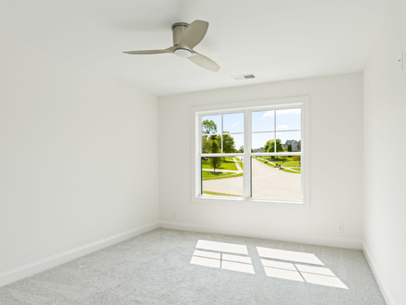 Empty bright bedroom with white walls, gray carpet, and a three-blade ceiling fan; window shows a sunny suburban street outside.