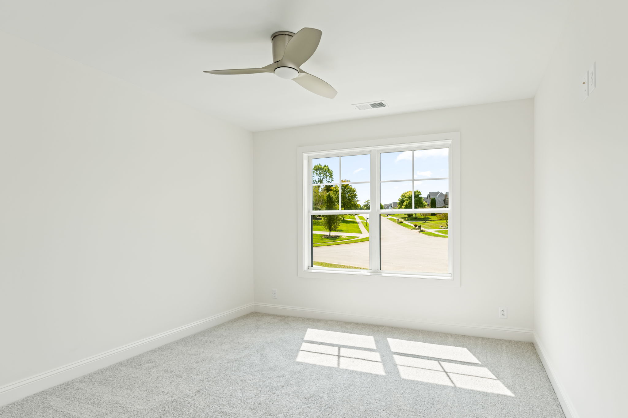 Empty bright bedroom with white walls, gray carpet, and a three-blade ceiling fan; window shows a sunny suburban street outside.