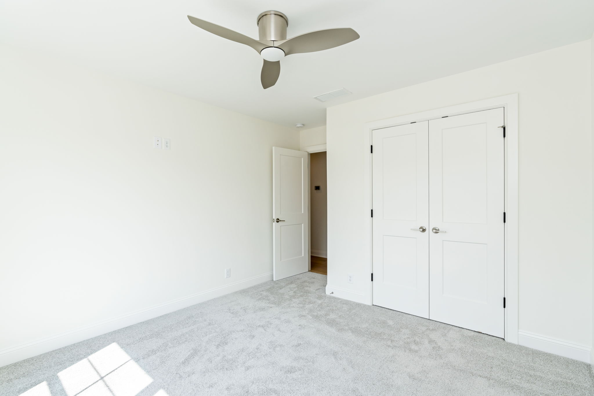 Bright empty bedroom with white walls, light carpet, a modern ceiling fan, an open doorway, and a large double-door closet