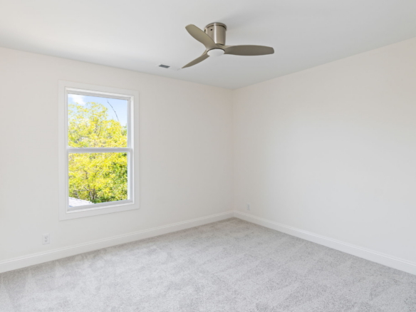 Bright empty bedroom with beige walls, light carpet, a window showing green trees, and a three-blade ceiling fan.