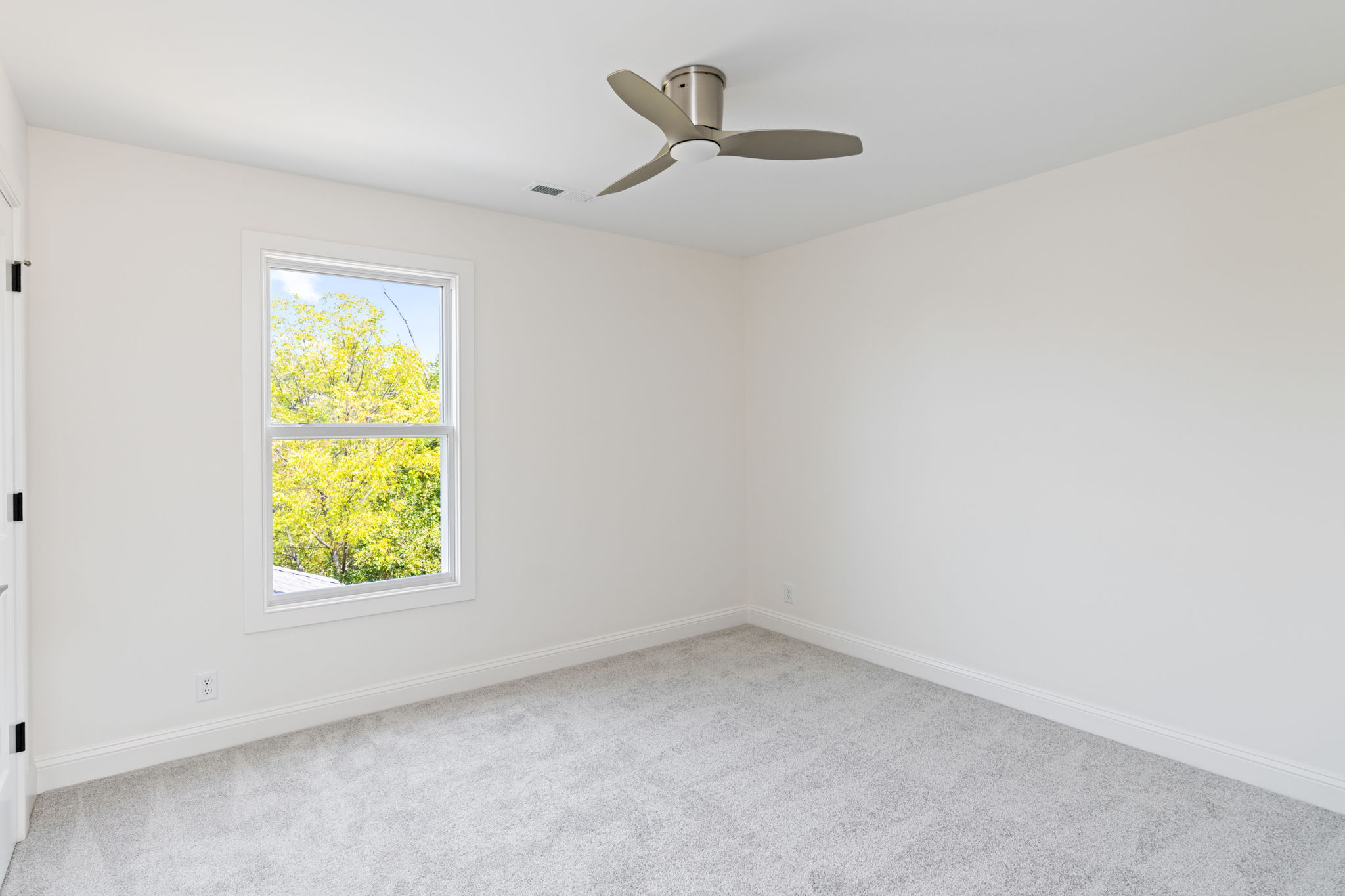 Bright empty bedroom with beige walls, light carpet, a window showing green trees, and a three-blade ceiling fan.