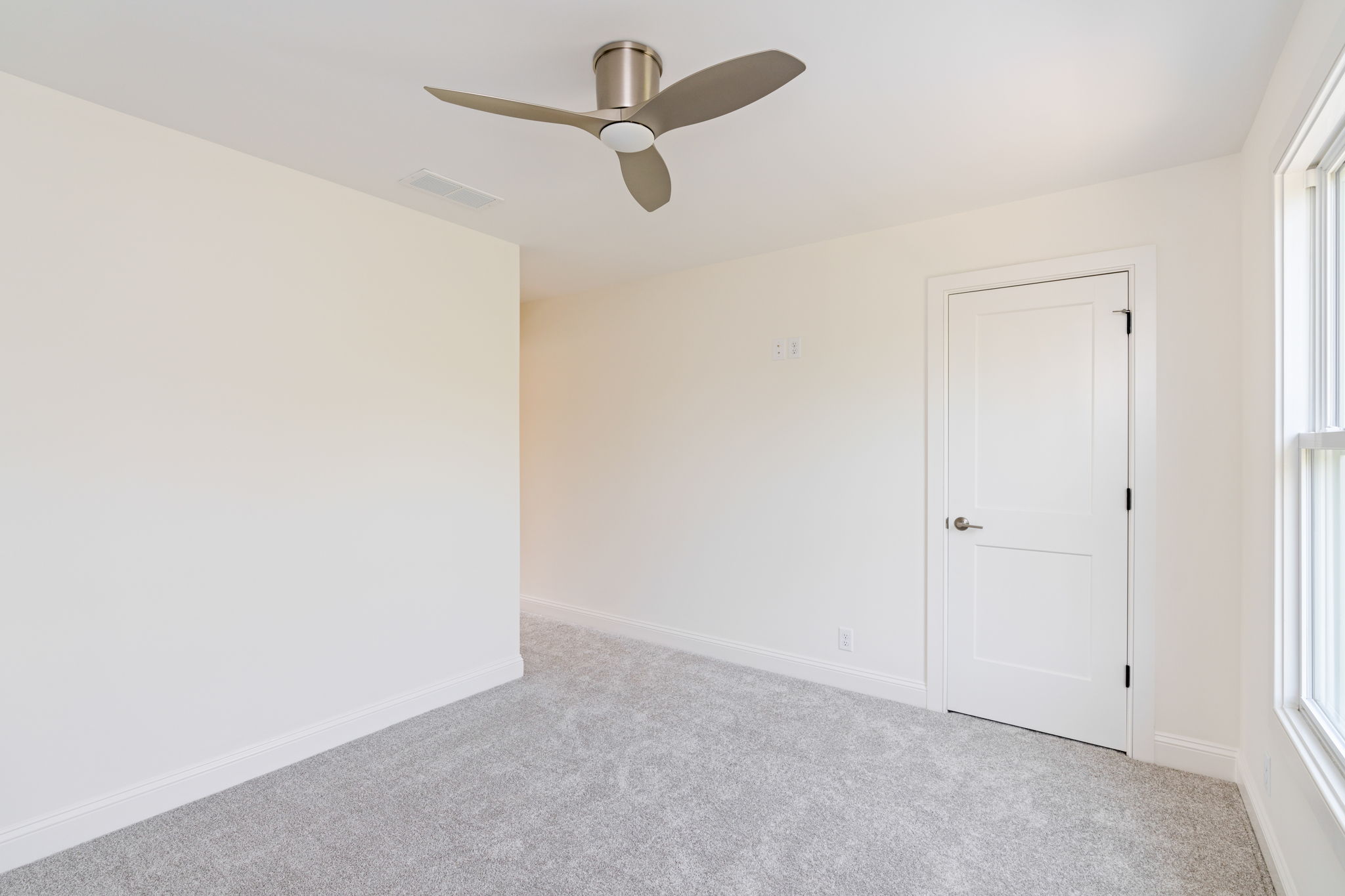 Empty light-filled room with white walls, gray carpet, a modern ceiling fan, a white paneled door, and a large window to the right.