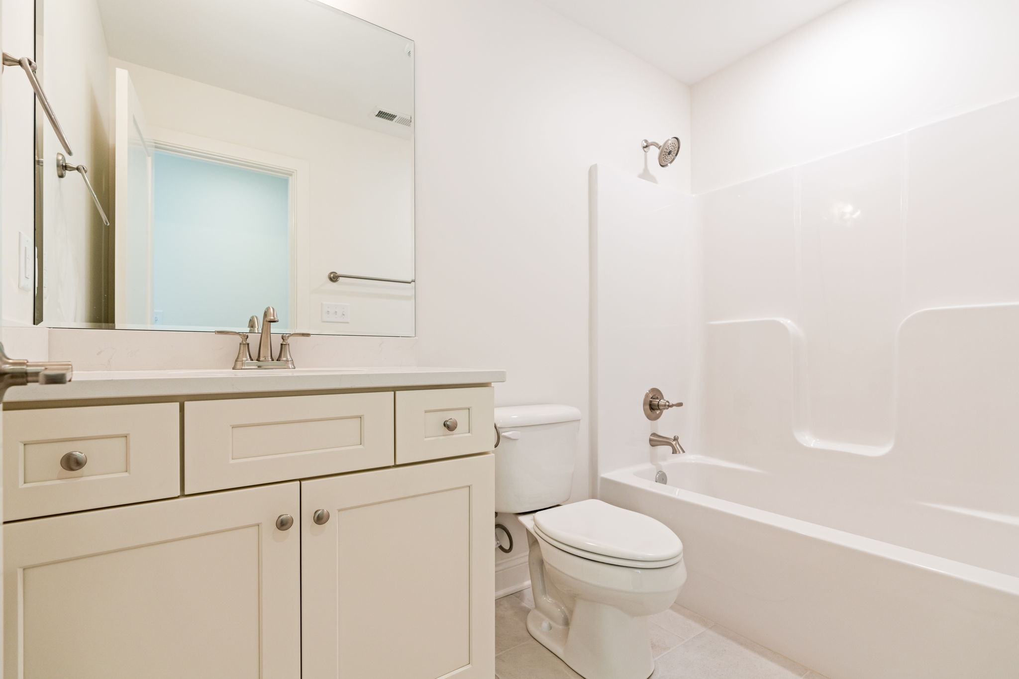 White bathroom with a vanity, large mirror, chrome faucet, and storage drawers next to a toilet and bathtub/shower area.