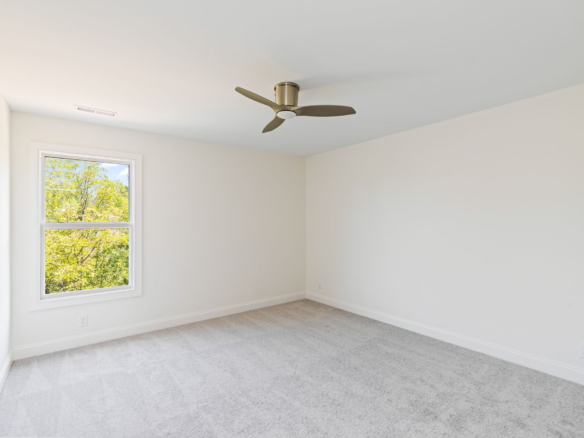 Empty, sunlit bedroom with beige walls, white trim, light carpet, a window showing green trees, and a modern ceiling fan in the center of the ceiling.
