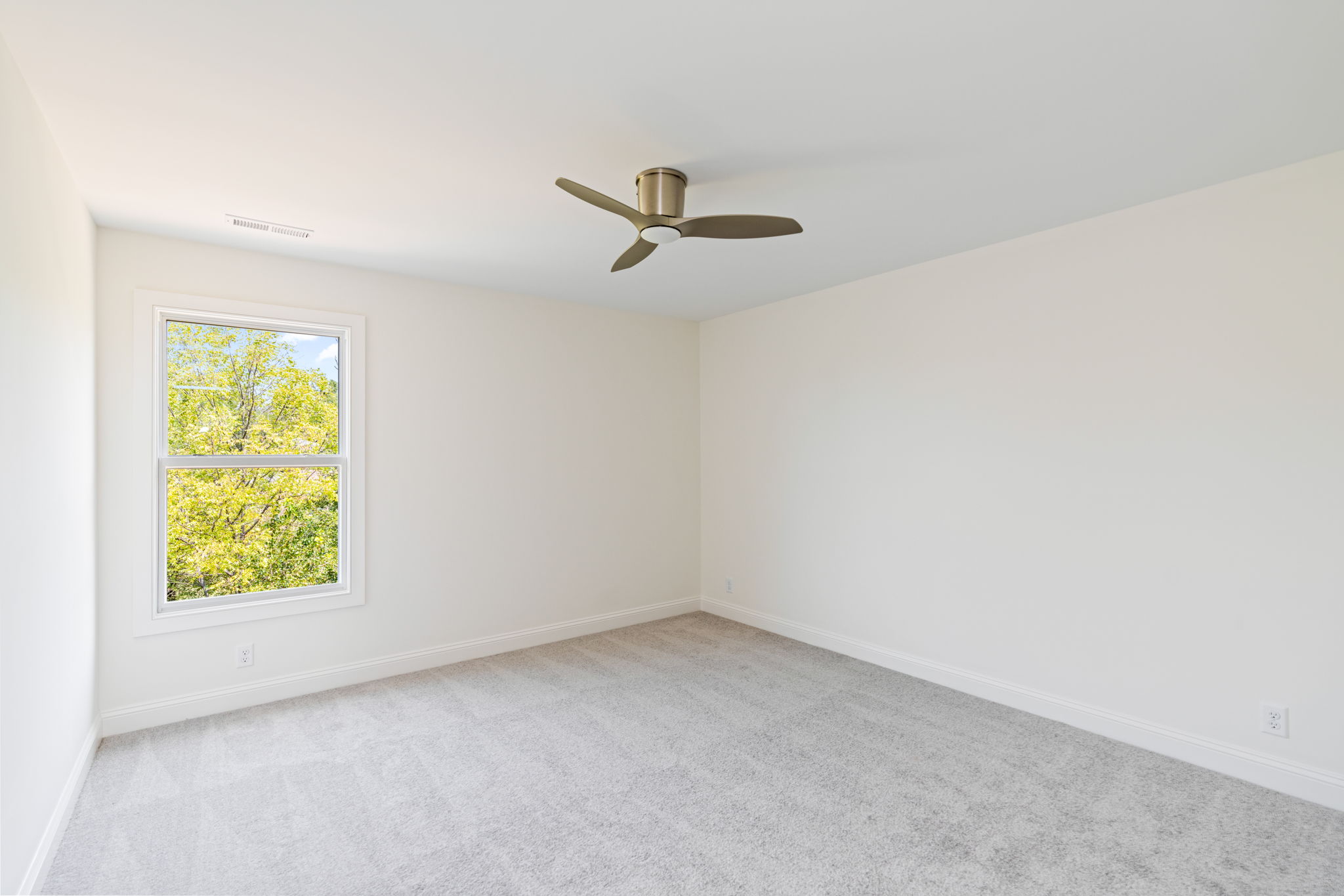 Empty, sunlit bedroom with beige walls, white trim, light carpet, a window showing green trees, and a modern ceiling fan in the center of the ceiling.