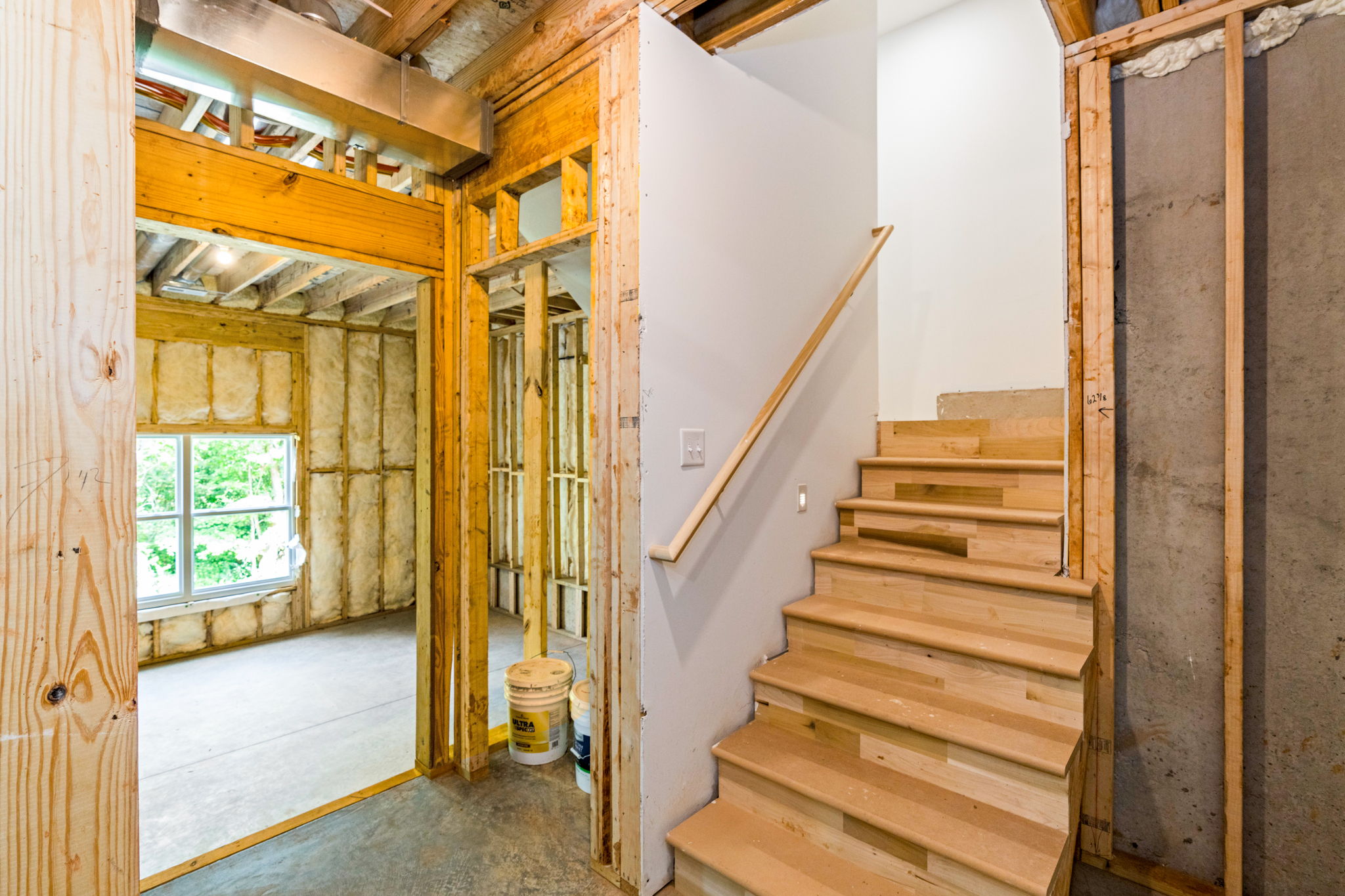 Under-construction interior with exposed wooden studs, insulation, and a wooden staircase on the right side, near a white wall column.