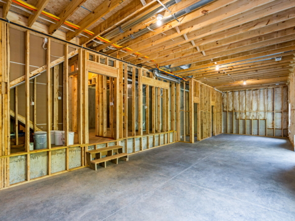 Interior of a house under construction with exposed wooden studs, ceiling joists, and utility pipes above.