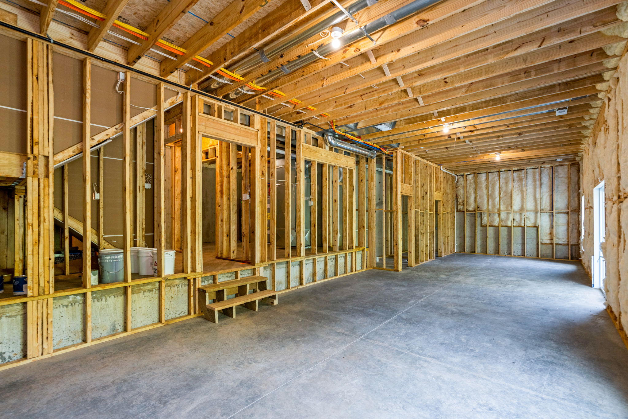Interior of a house under construction with exposed wooden studs, ceiling joists, and utility pipes above.