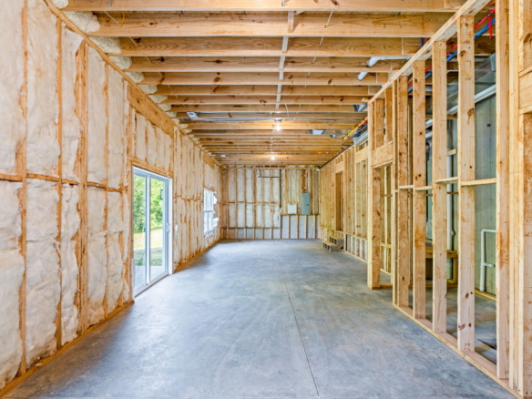 Unfinished interior of a house with exposed wooden studs, fiberglass insulation, and visible plumbing along the ceiling beams.