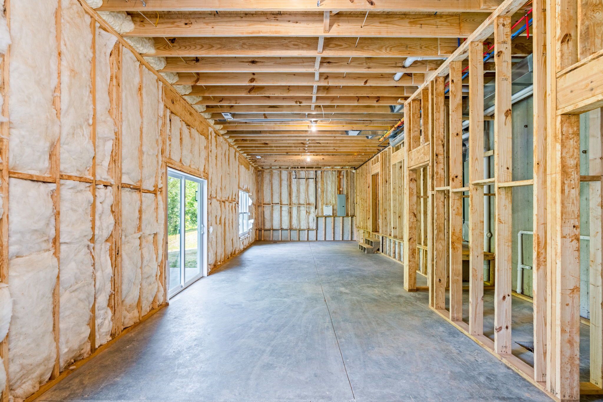 Unfinished interior of a house with exposed wooden studs, fiberglass insulation, and visible plumbing along the ceiling beams.