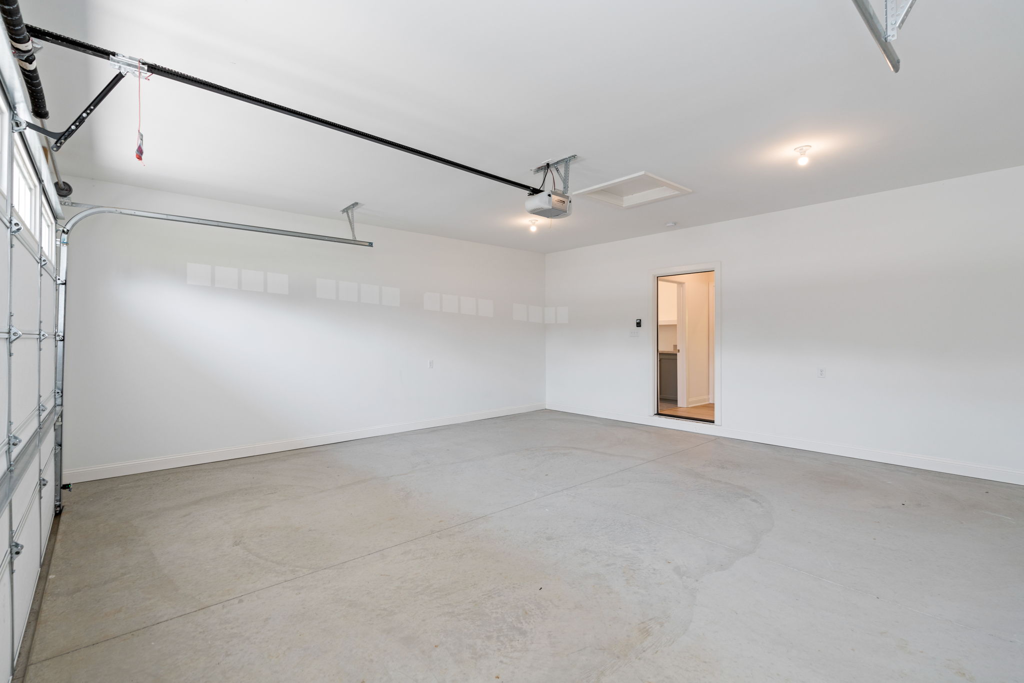 Empty two-car garage with white walls and a concrete floor, ceiling-mounted garage door opener and tracks along the ceiling, and a door to the house on the right.