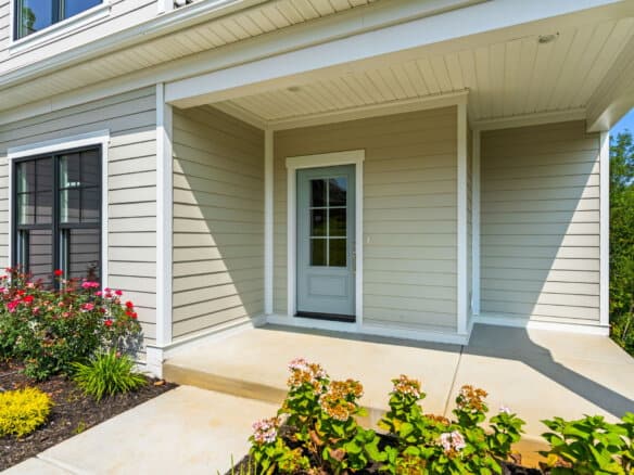 Front porch with white door and beige siding, surrounded by colorful flowers.