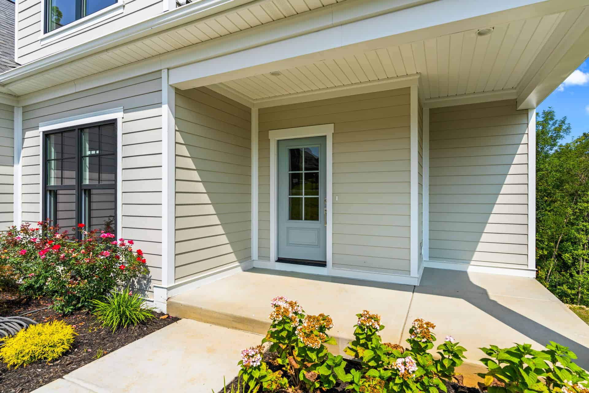 Front porch with white door and beige siding, surrounded by colorful flowers.