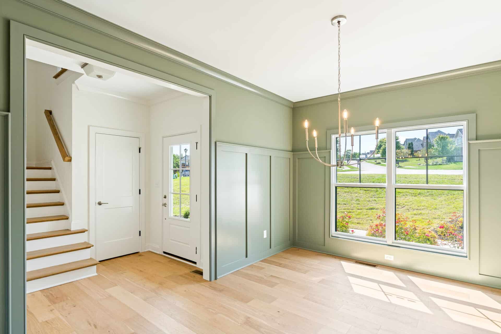 Bright foyer with sage green walls, a hanging chandelier, and white trim; stairs rise on the left and a large window looks out to a suburban street.