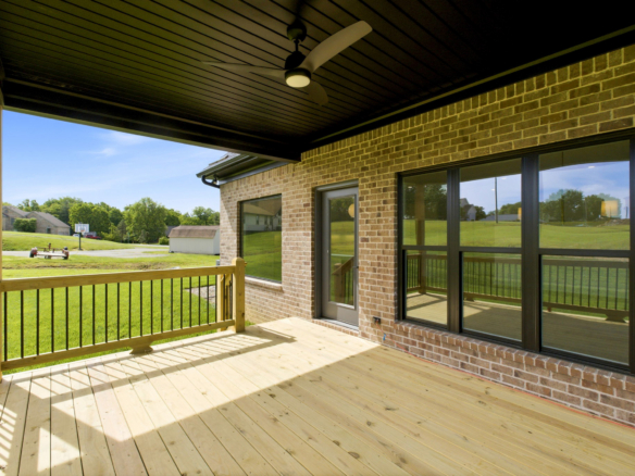 Covered wooden deck attached to a brick house, with a ceiling fan and large glass doors overlooking a green backyard.