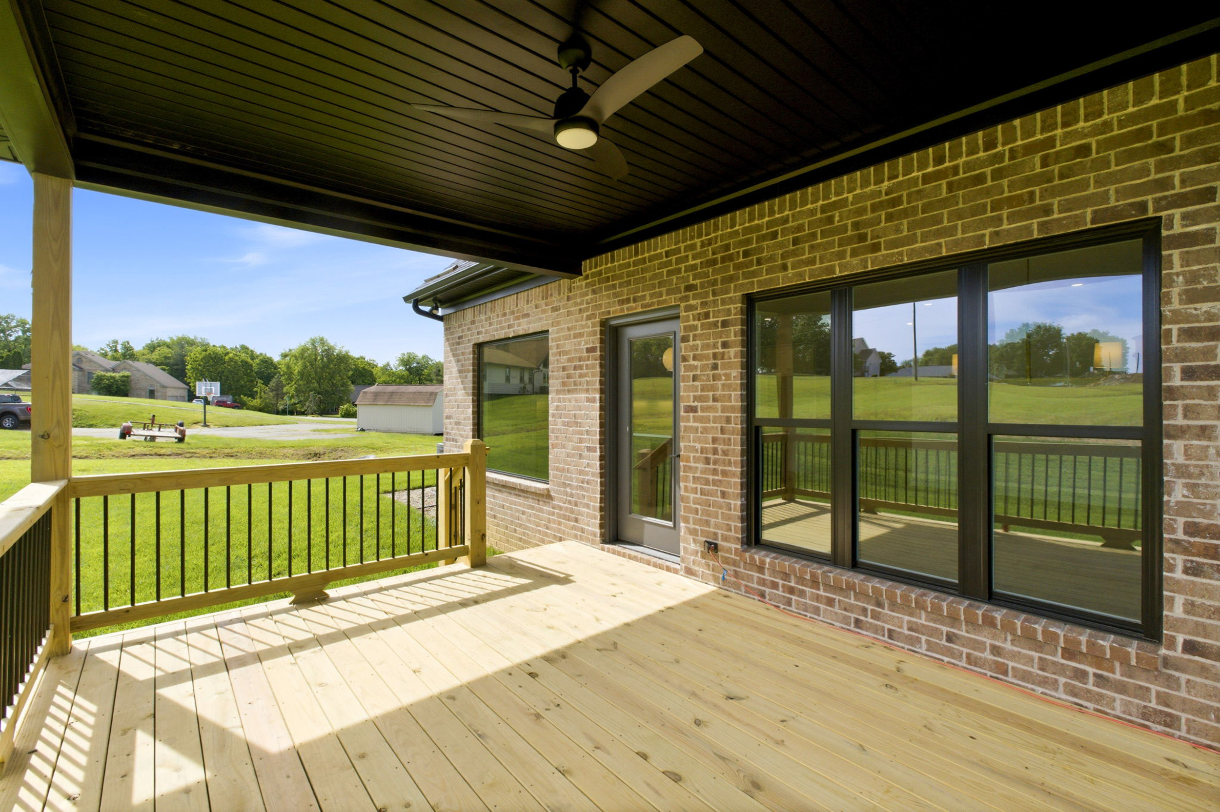Covered wooden deck attached to a brick house, with a ceiling fan and large glass doors overlooking a green backyard.