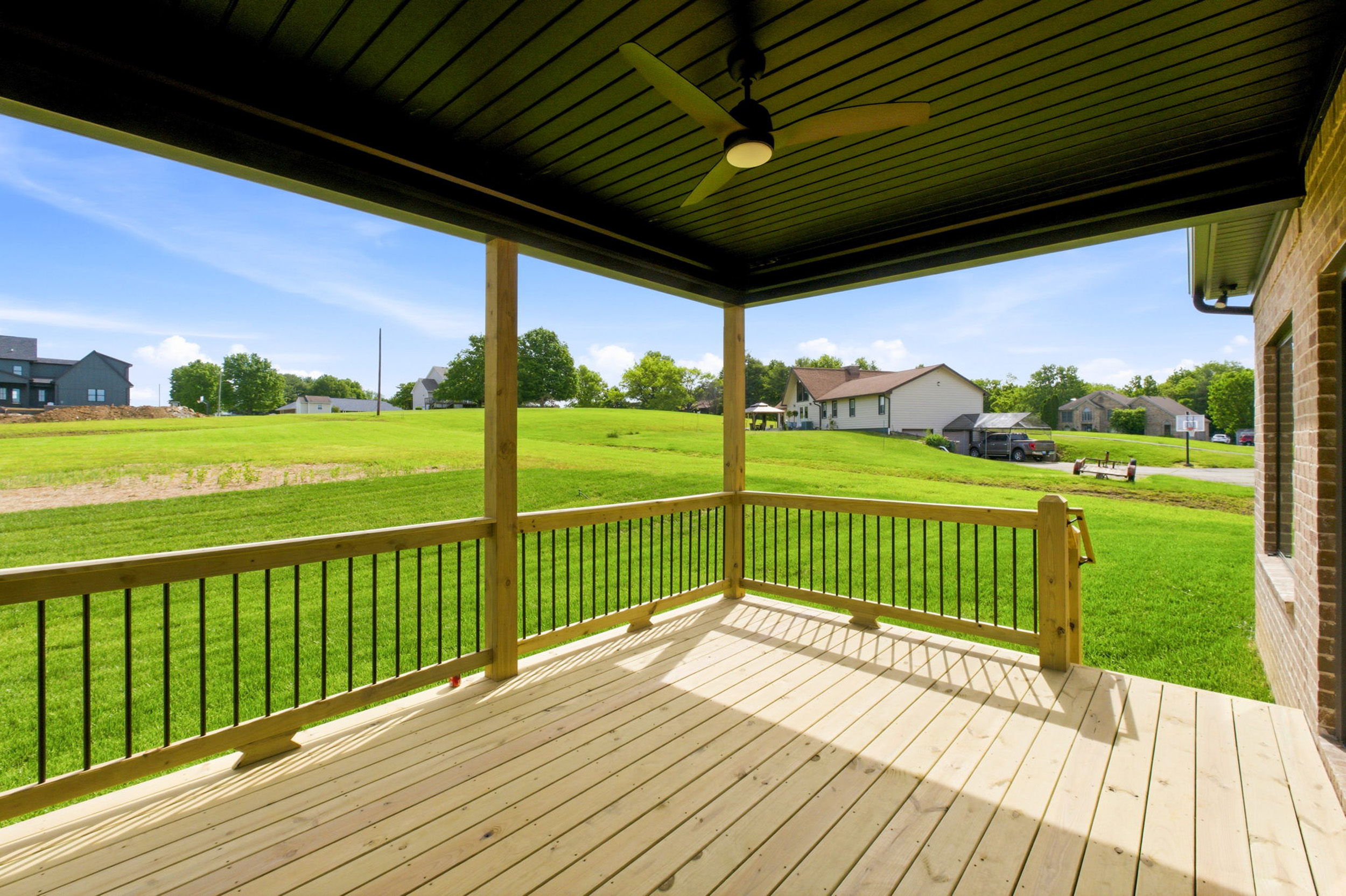 Wooden deck under a dark ceiling with a ceiling fan, overlooking a green suburban yard and houses in the distance.