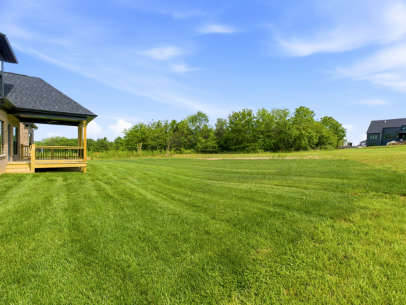Brick house with a wooden porch on the left, a large grassy lawn, and a line of trees under a clear blue sky.