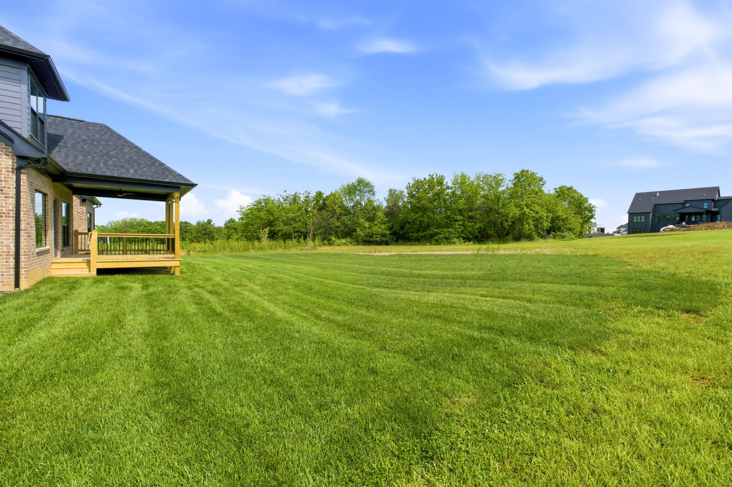 Brick house with a wooden porch on the left, a large grassy lawn, and a line of trees under a clear blue sky.
