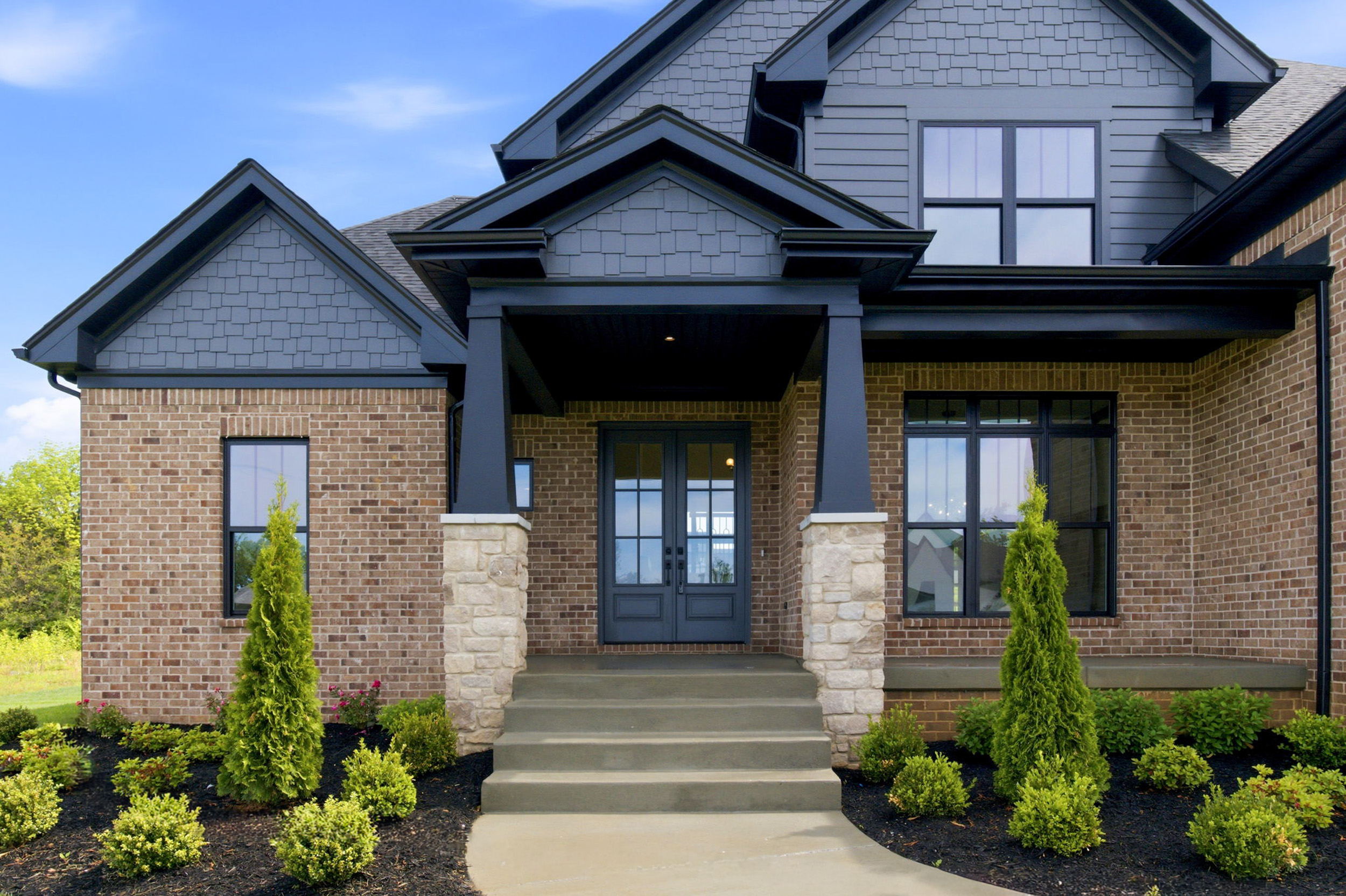 Front porch of a brick house with grey siding, black-framed windows, and stone columns supporting a covered entryway.