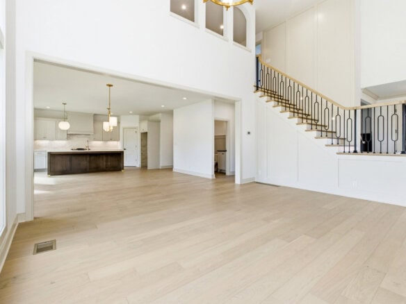 Bright open-concept living area with light wood floors, white walls, a kitchen to the left, and a curved staircase with black balusters nearby.