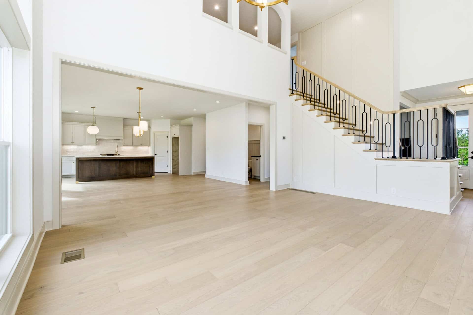 Bright open-concept living area with light wood floors, white walls, a kitchen to the left, and a curved staircase with black balusters nearby.