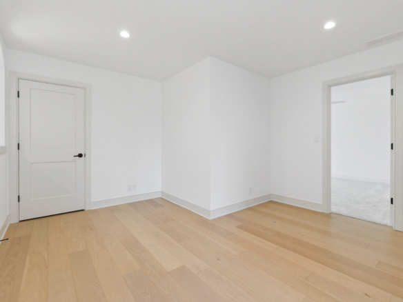 Empty white room with light wood floors, a closed door on the left and an open doorway to another room on the right, ceiling lights visible.