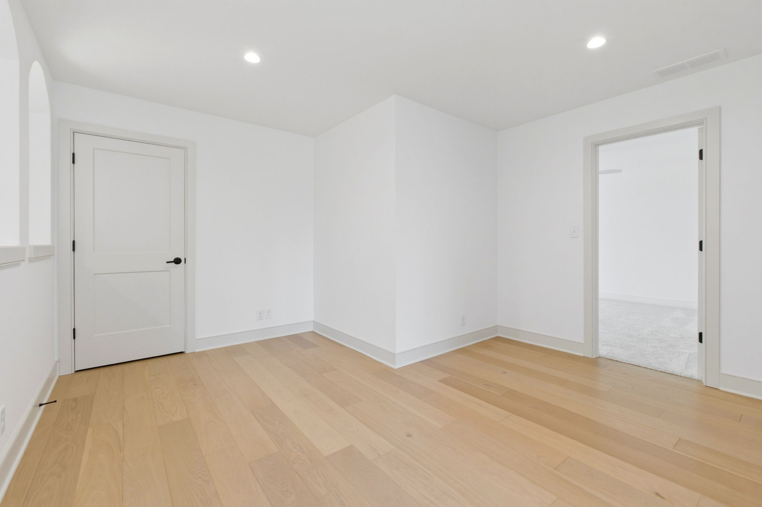 Empty white room with light wood floors, a closed door on the left and an open doorway to another room on the right, ceiling lights visible.