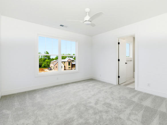 Bright empty room with white walls, a large window showing a suburban street, and a white ceiling fan above.
