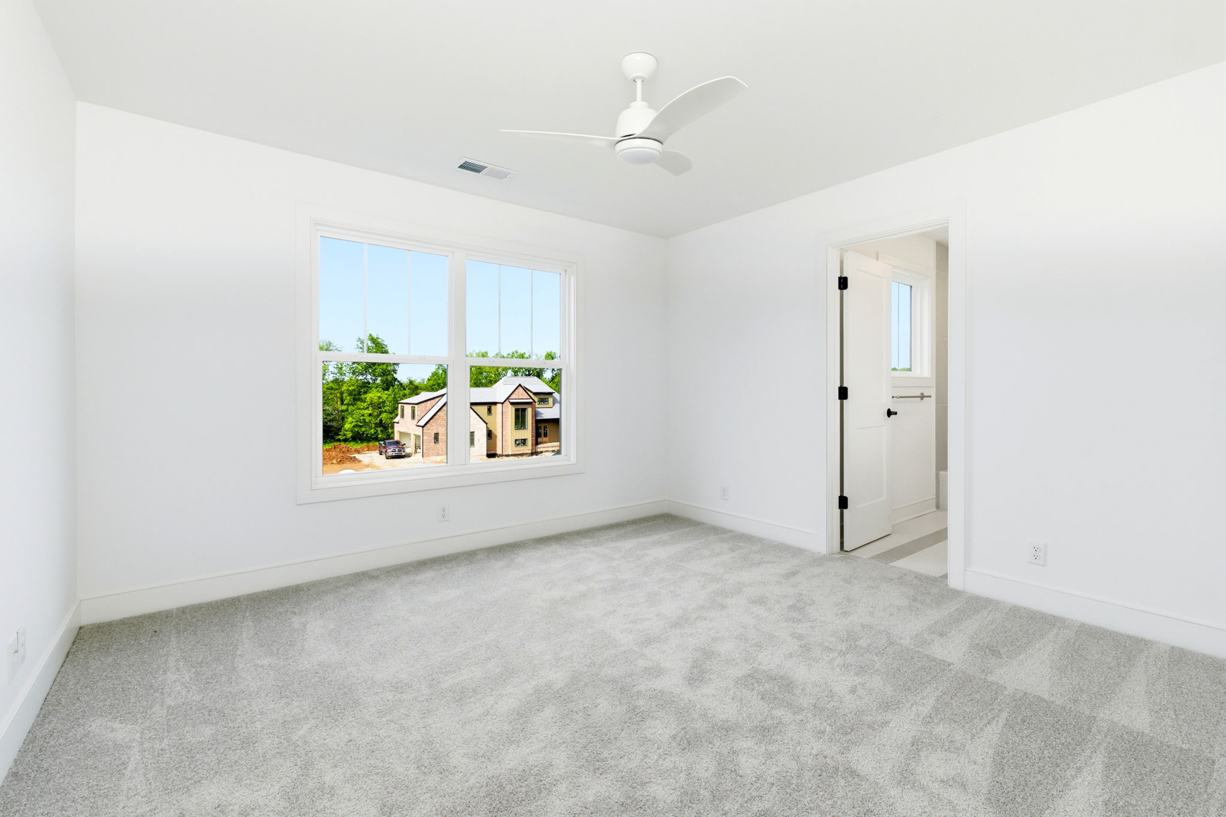 Bright empty room with white walls, a large window showing a suburban street, and a white ceiling fan above.