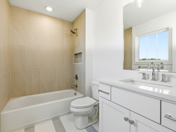 Bright white bathroom with a bathtub and beige tiled shower surround, and a white vanity beneath a window. (Informative)