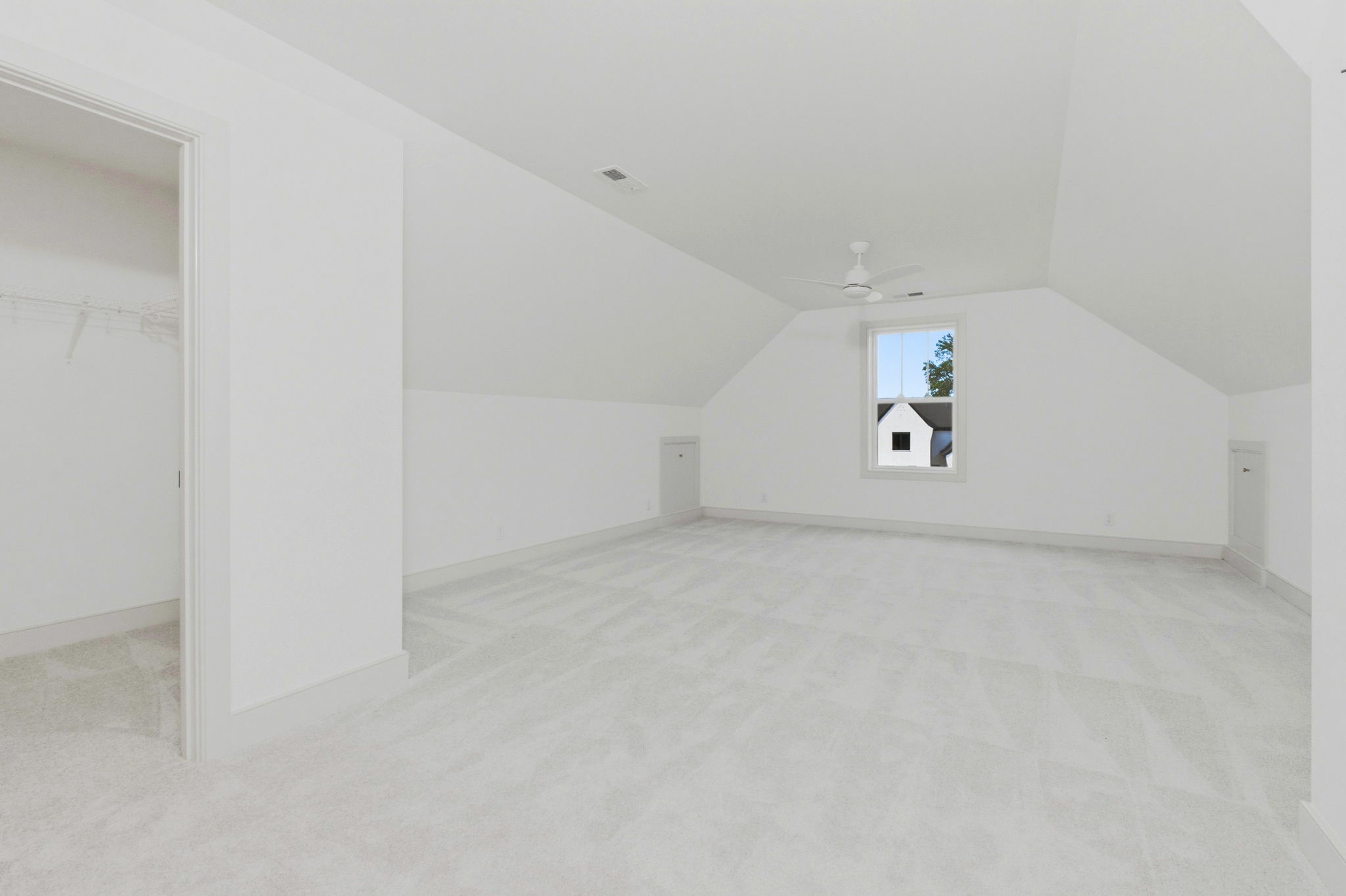 Empty white room with a vaulted ceiling, ceiling fan, and a small window showing a house outside on the right side of the room.