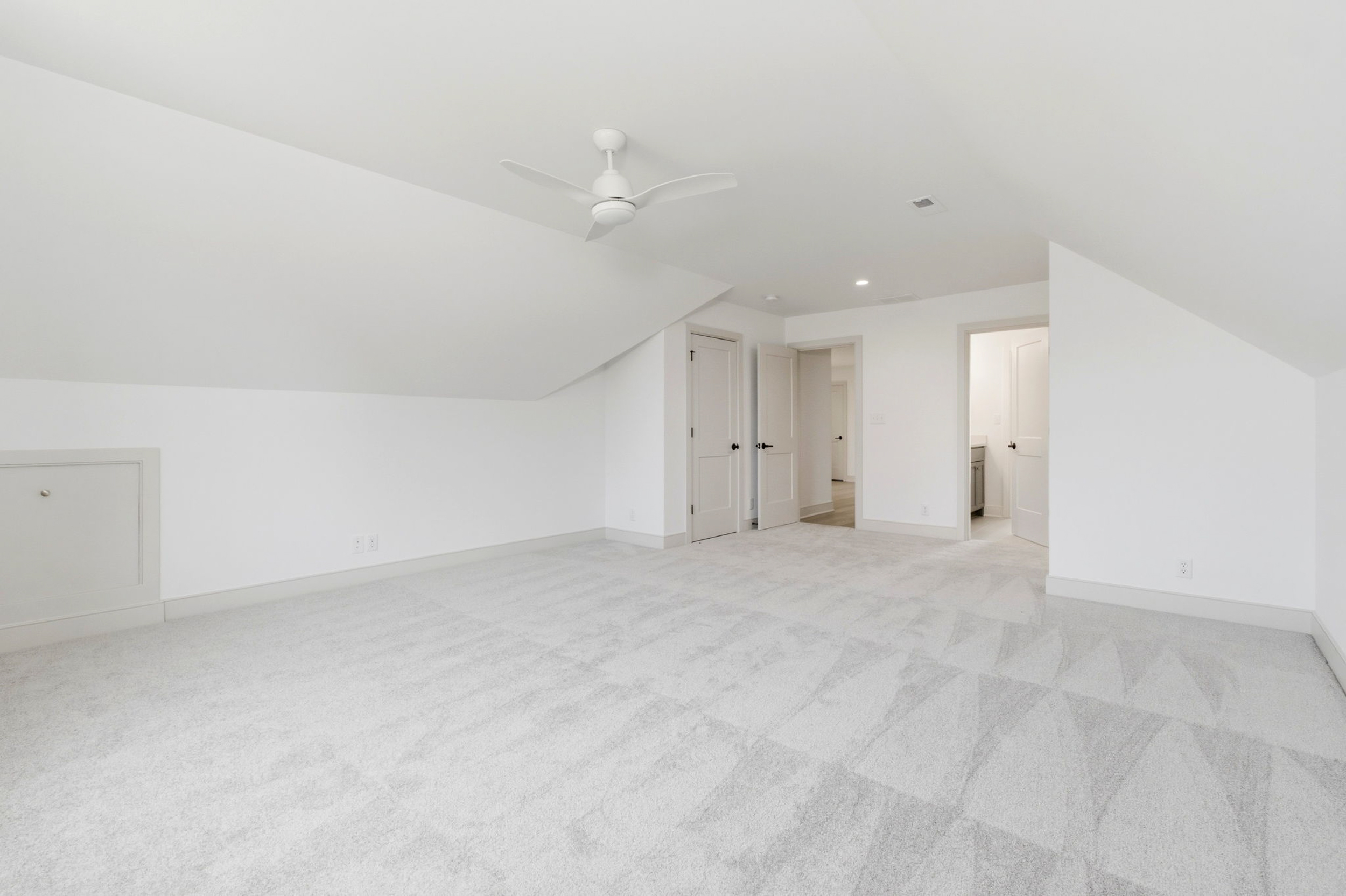 Bright empty attic bedroom with white walls and a white ceiling fan, open closet/doorways visible in the background.