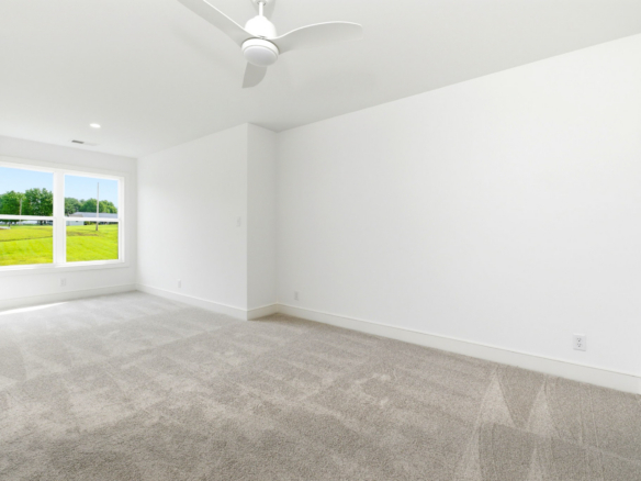 Bright empty room with white walls, light carpet, ceiling fan, and a large window looking onto a green lawn.