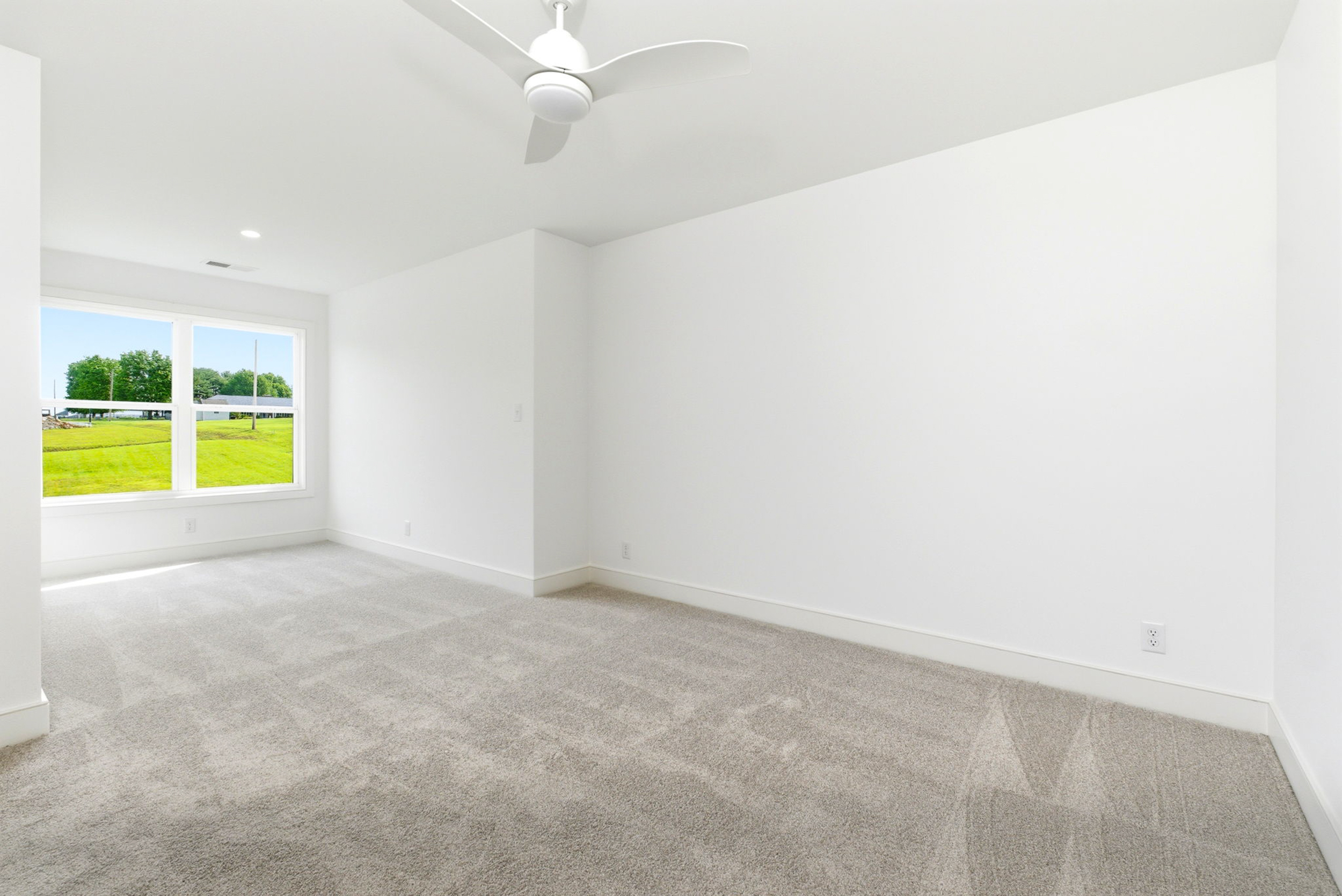 Bright empty room with white walls, light carpet, ceiling fan, and a large window looking onto a green lawn.