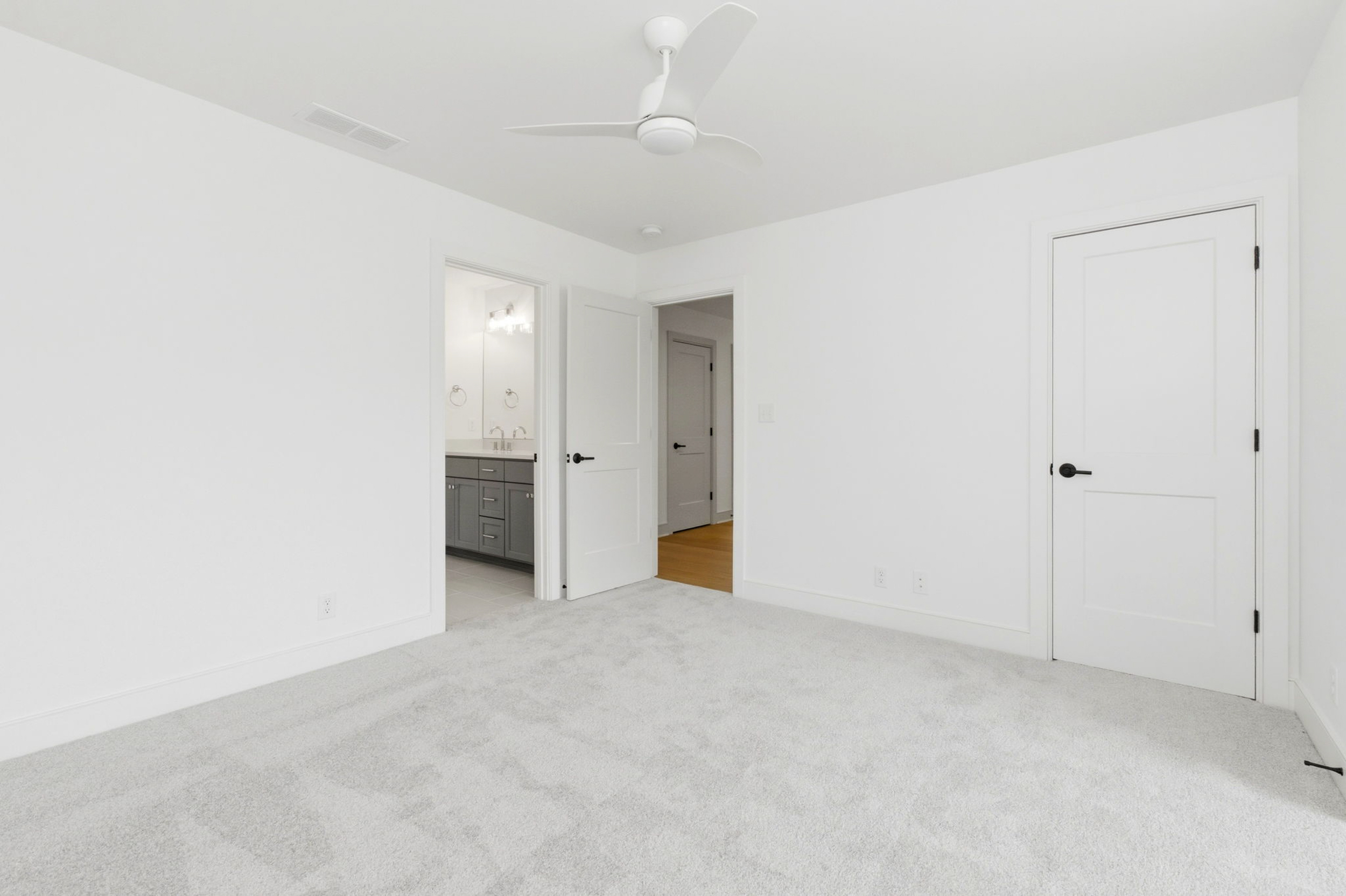 Bright white bedroom with two open doors; bathroom with grey vanity visible on the left and a white ceiling fan overhead.