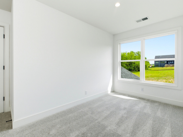 Bright empty white room with a large window letting in sunlight; green trees and a house visible outside.