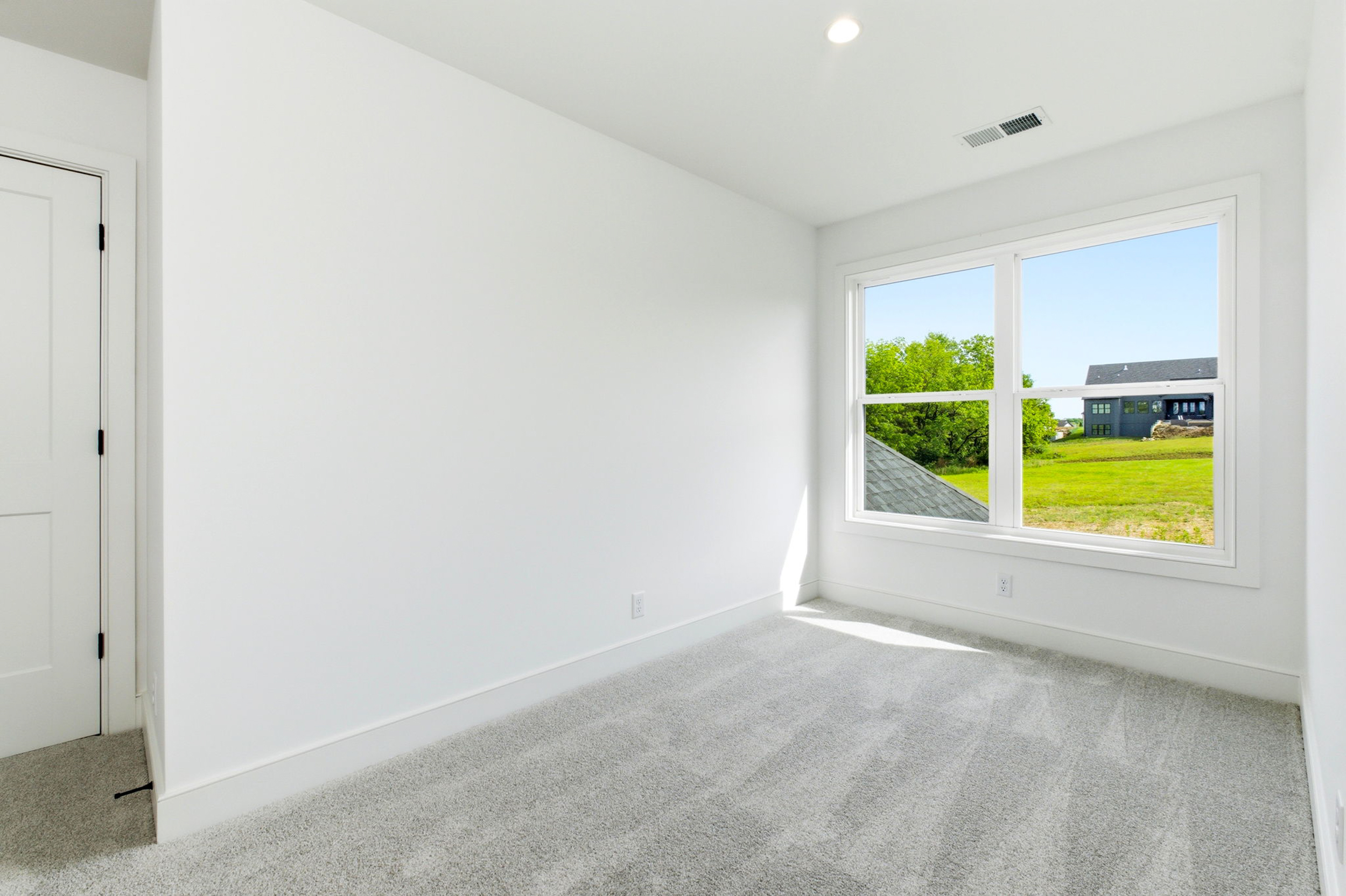 Bright empty white room with a large window letting in sunlight; green trees and a house visible outside.