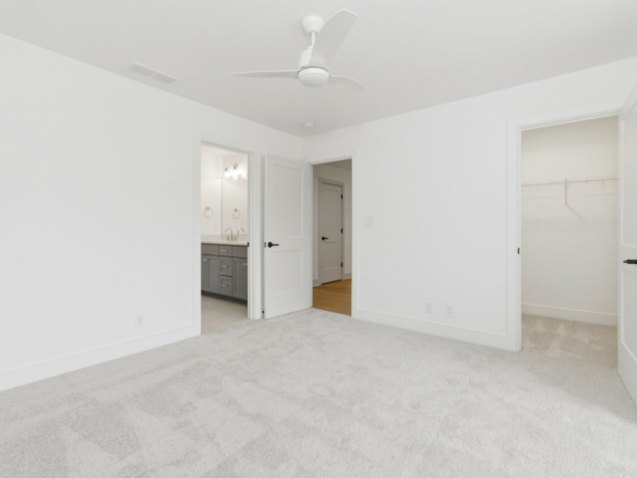 Empty white bedroom with light gray carpet, ceiling fan, and open doors to a bathroom and closet.