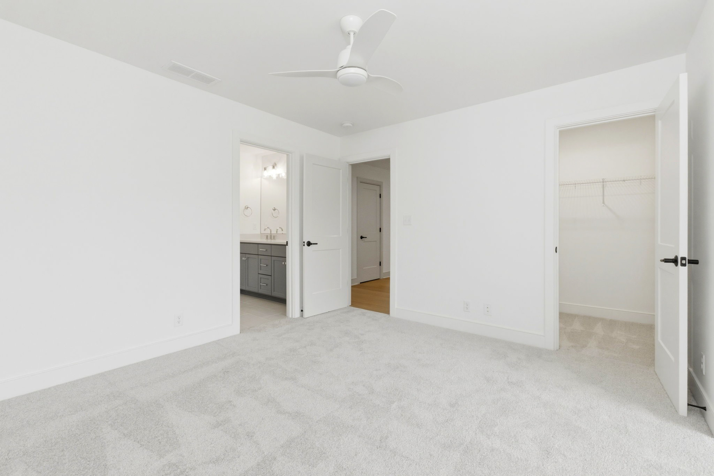 Empty white bedroom with light gray carpet, ceiling fan, and open doors to a bathroom and closet.
